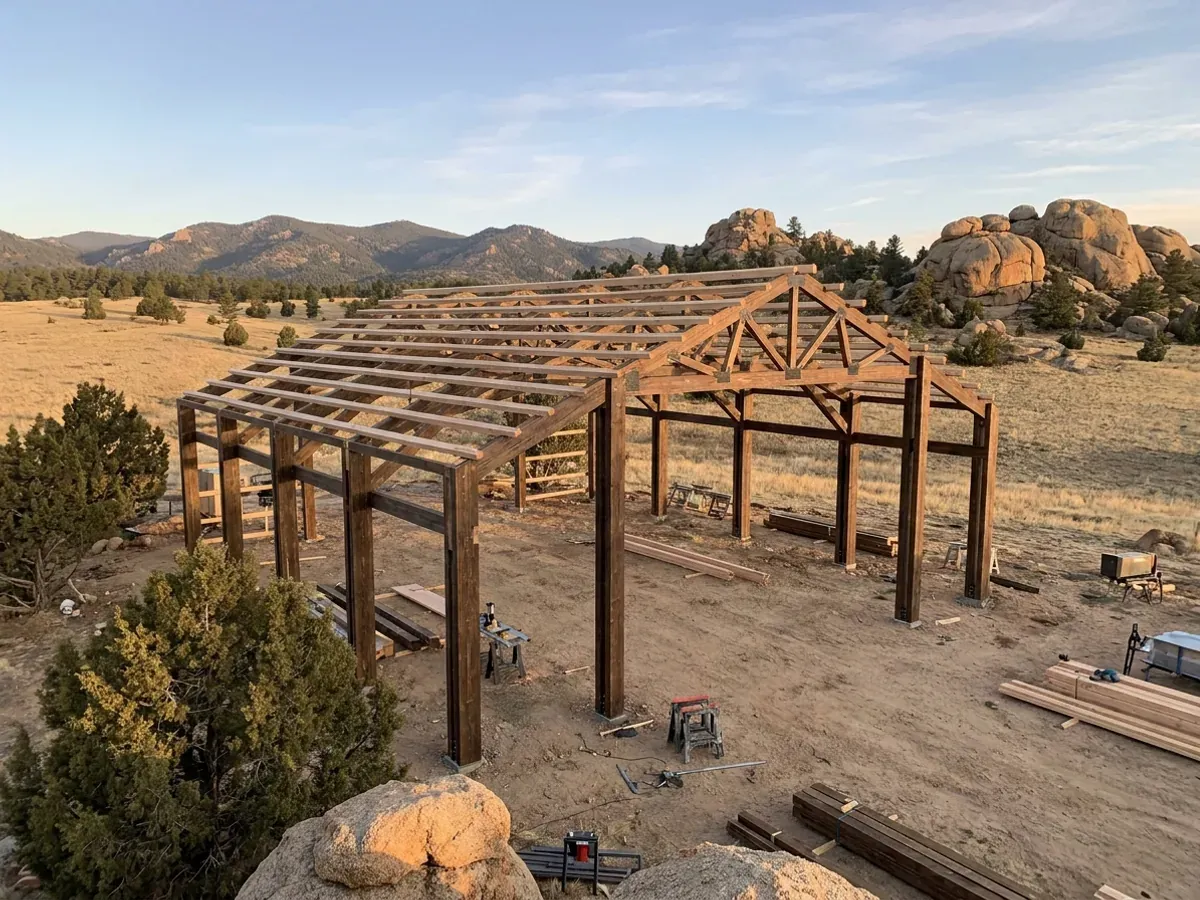 A pole barn home under construction on open grassland among granite boulders, with heavy timber columns and wide-span roof trusses.