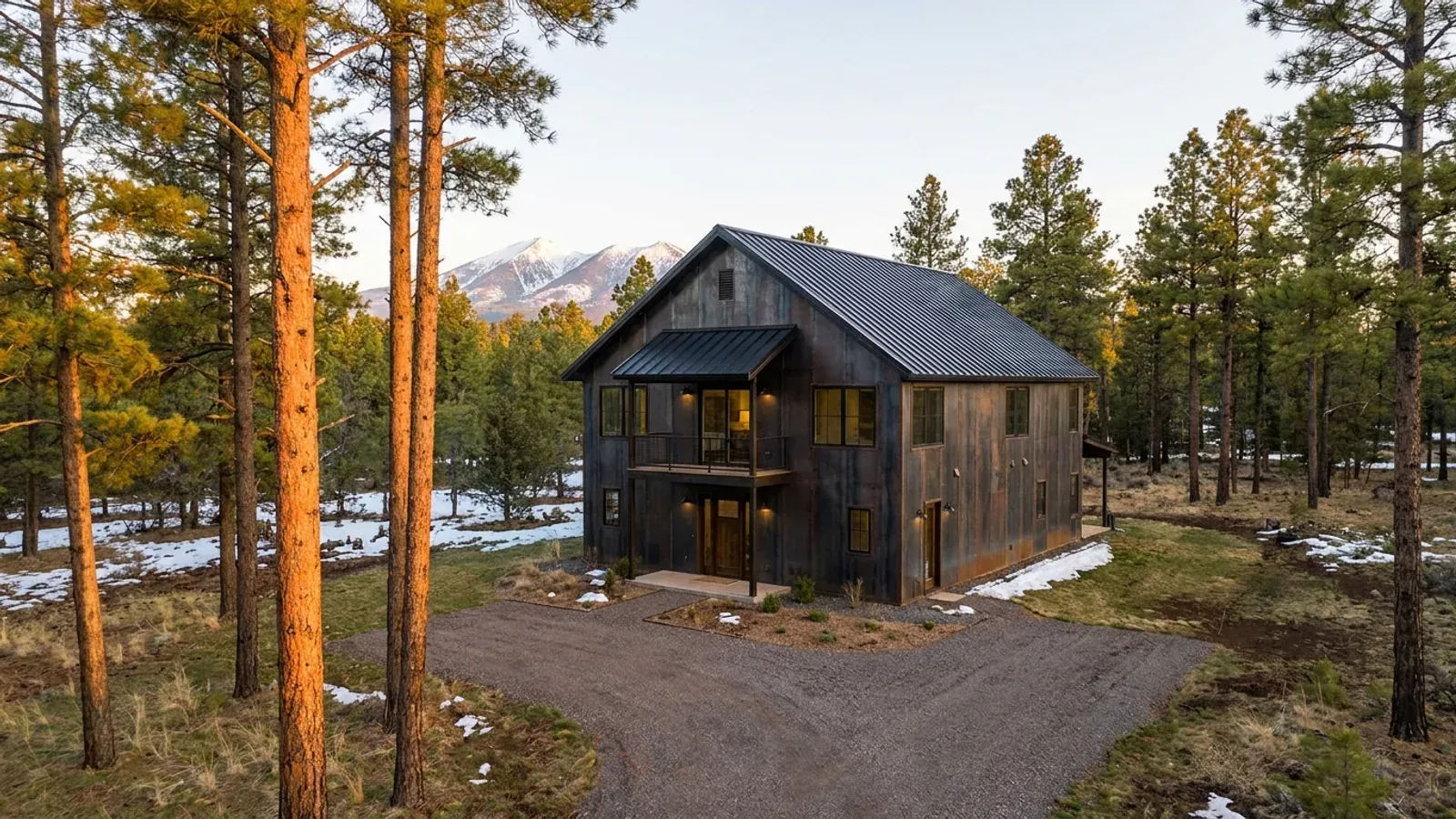 A completed two-story barndominium among ponderosa pines, with second-story windows, a covered balcony and snow-capped peaks beyond.
